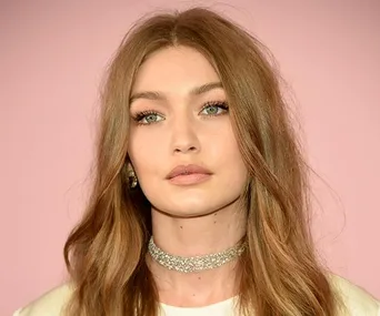 A woman with long, light brown hair, wearing a sparkly choker necklace, against a soft pink background.