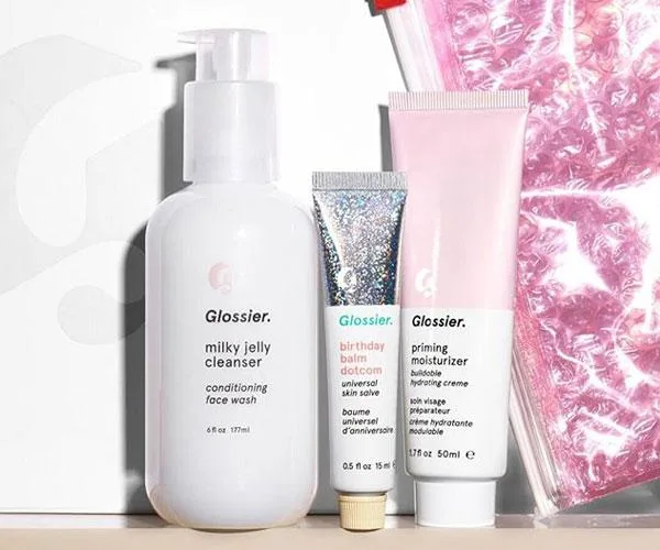 Glossier products including Milky Jelly Cleanser, Birthday Balm Dotcom, and Priming Moisturizer with pink pouch in background.