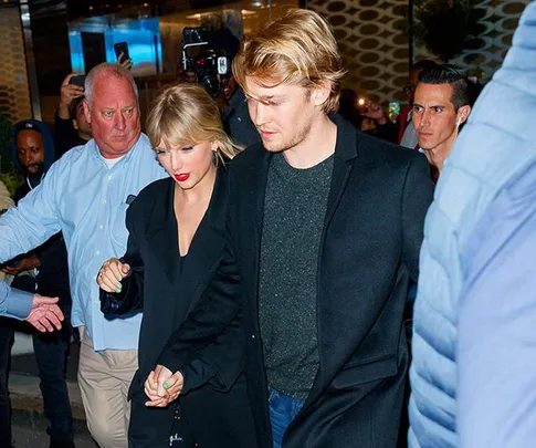 A couple holding hands and walking with security guards in a crowd at an evening event.