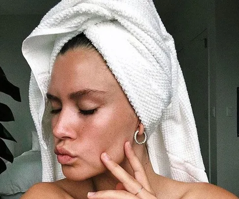 Woman with a towel on her head, eyes closed, wearing hoop earrings and touching her cheek with a peaceful expression.