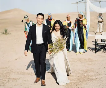 A couple walks in the desert in wedding attire, followed by men in colorful robes, with a tent setup in the background.