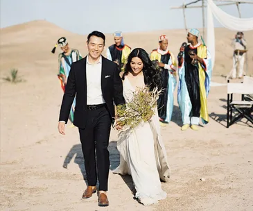 A couple walks in the desert in wedding attire, followed by men in colorful robes, with a tent setup in the background.