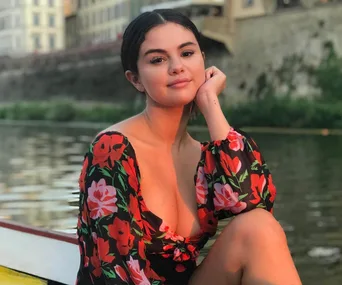 Young woman smiling in floral dress, sitting by a riverside with blurred buildings in the background.