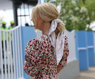 A woman with blonde hair in a low bun, wearing a floral dress and a scarf, standing outdoors near a blue fence.