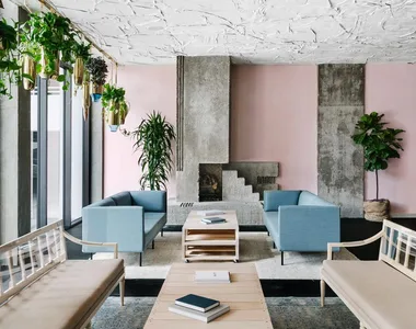 Modern lounge with pastel walls, blue sofas, a central fireplace, plants hanging from ceiling and a wooden coffee table.