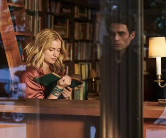 A woman reads a book in a cozy bookstore; a man stands behind her, partially obscured by a glass reflection.