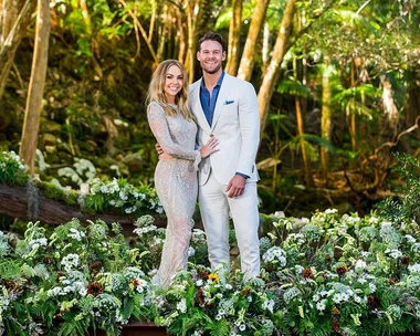 Man and woman in formal attire standing among lush greenery and flowers in a forest setting.