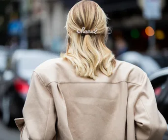 Woman with blond hair, wearing a beige coat and a Chanel hair clip, seen from the back on a city street.