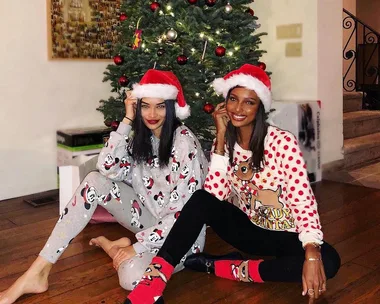 Two women in Christmas pajamas and Santa hats sitting by a decorated tree, smiling at the camera.