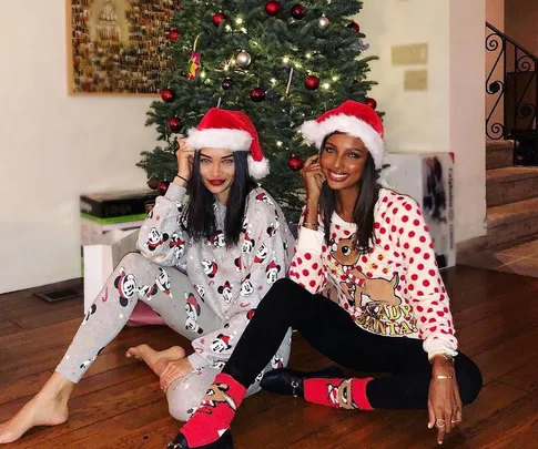 Two women in Christmas pajamas and Santa hats sitting by a decorated tree, smiling at the camera.