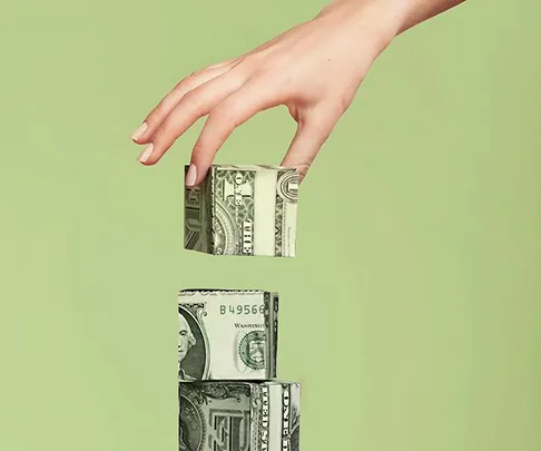 A hand stacking cube-shaped blocks of dollar bills against a green background.