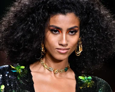 A woman with voluminous curly hair, bold eye makeup, gold jewelry, and a sequined outfit on a fashion runway.