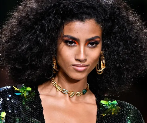 A woman with voluminous curly hair, bold eye makeup, gold jewelry, and a sequined outfit on a fashion runway.