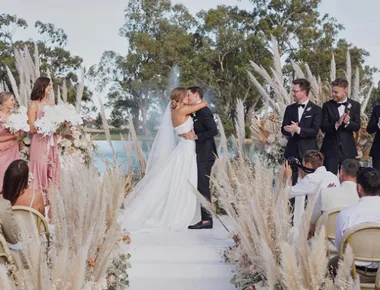 Bride and groom kiss at outdoor wedding ceremony surrounded by pampas grass, bridesmaids in pink, and groomsmen in black.