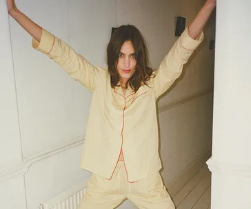 Woman in beige pajamas with red piping, standing in a hallway with arms raised, smiling playfully at the camera.