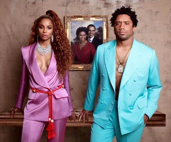 A couple in vibrant suits pose next to a portrait of Barack and Michelle Obama on a wall.