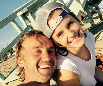 Two people smiling, taking a selfie at the beach on a sunny day.