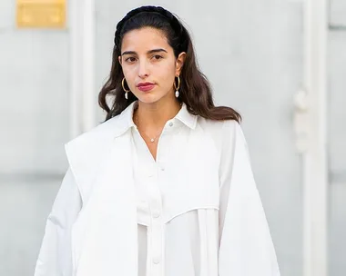 A woman wearing a white shirt with statement earrings, standing against a light gray background.