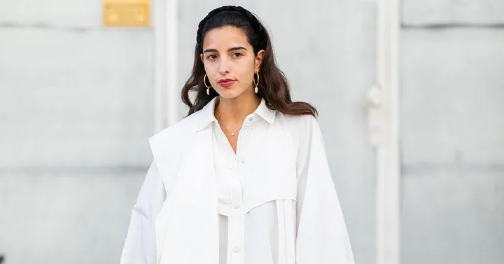 A woman wearing a white shirt with statement earrings, standing against a light gray background.