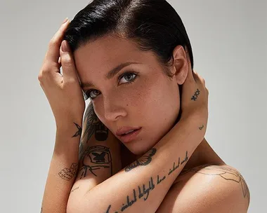 A person with tattoos poses with hands on head, looking directly at the camera, short dark hair slicked back.