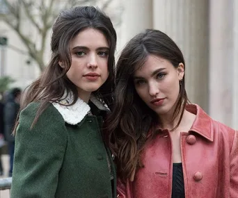 Two women with long hair, wearing green and pink coats, standing closely together outdoors.