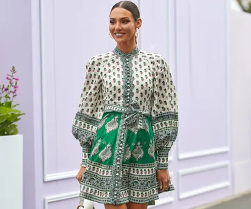 Woman in a patterned green and white dress smiling, with a background of light purple walls and flowers.