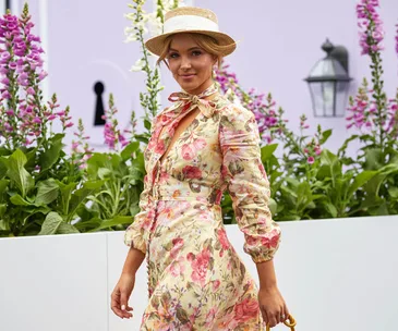 Woman in a floral dress and straw hat walking beside a garden with pink and white flowers.