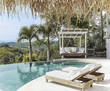 Luxury villa poolside with loungers, canopy bed, palm trees, scenic hills, and a clear sky in Australia.