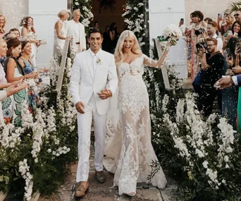 Smiling bride in lace gown and groom in white suit walk down flower-lined aisle as guests cheer and take photos.