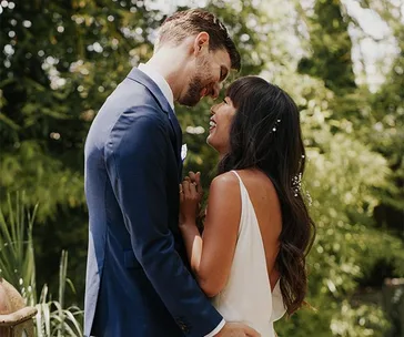 A couple in wedding attire embraces and laughs under a lush green tree canopy.