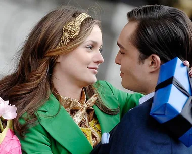 A couple embraces, smiling, with the man holding a blue gift box wrapped in black ribbon.