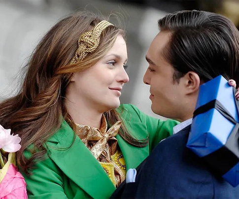 A couple embraces, smiling, with the man holding a blue gift box wrapped in black ribbon.