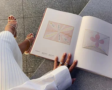 Person sitting with a book open on their lap, showing abstract illustrations, wearing white sandals and skirt.