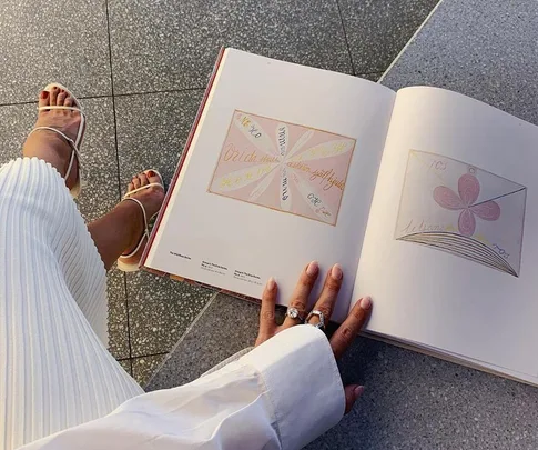 Person sitting with a book open on their lap, showing abstract illustrations, wearing white sandals and skirt.