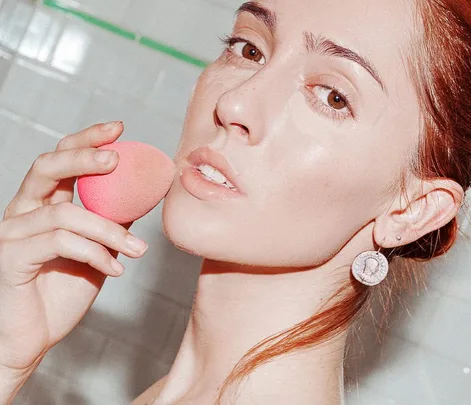 A woman applying makeup with a pink makeup sponge in a bathroom setting.