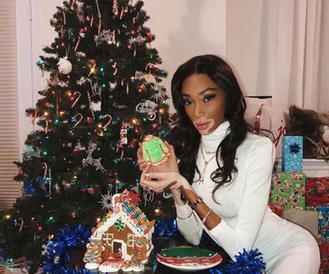 Woman in white holding decorated cookie by Christmas tree and gingerbread house.