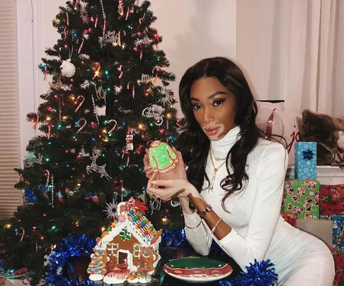 Woman in white holding decorated cookie by Christmas tree and gingerbread house.