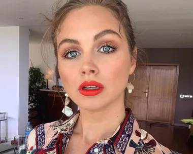 A person with bright blue eyes and red lipstick, wearing a patterned shirt, poses indoors in front of wooden doors.