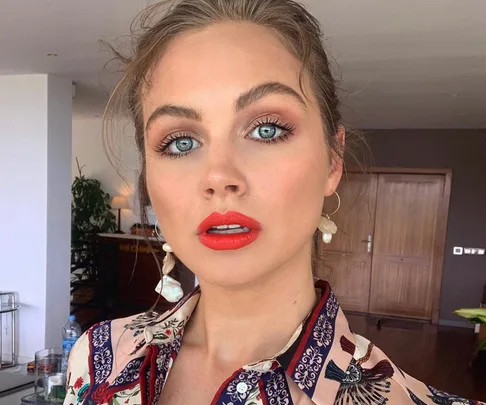 A person with bright blue eyes and red lipstick, wearing a patterned shirt, poses indoors in front of wooden doors.