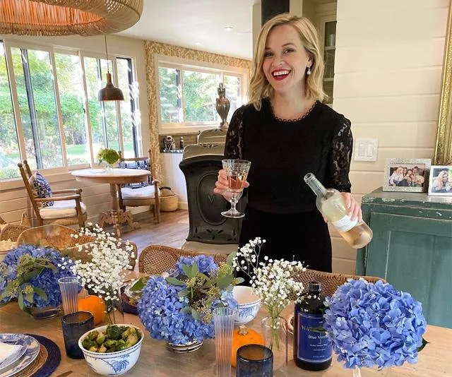 A woman in a black dress smiles, holding a wine glass and bottle, with a table of flowers and a cozy kitchen setting.