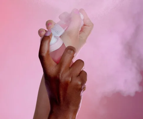 Hands spraying a mist against a pink background.