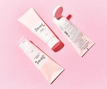 Three tubes of Korean exfoliator on a pink background, one with its cap open.