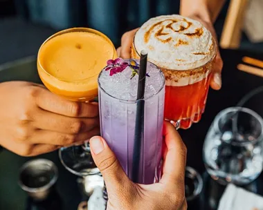 Hands holding three colorful cocktails, including one purple with a flower garnish.