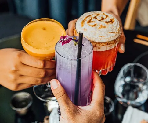 Hands holding three colorful cocktails, including one purple with a flower garnish.