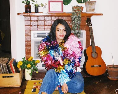 A person wearing a colorful, tinsel-like shawl sits near a fireplace, with a guitar and plants in the background.