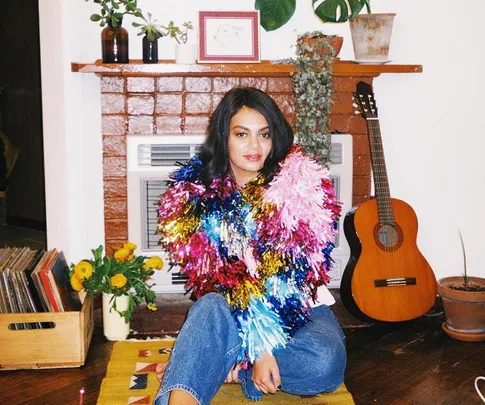 A person wearing a colorful, tinsel-like shawl sits near a fireplace, with a guitar and plants in the background.