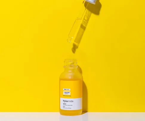 A dropper above a Hylamide Glow Radiance Booster bottle on a bright yellow background.