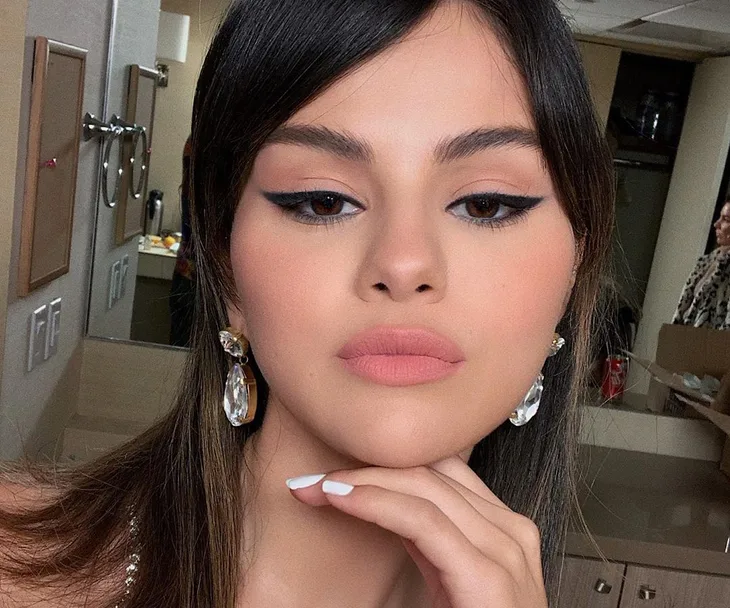 Selena Gomez with winged eyeliner and crystal earrings, posing in a makeup room.