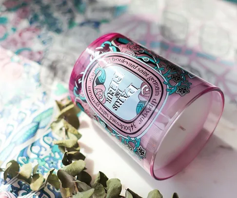 Pink Diptyque candle with ornate label, surrounded by patterned paper and a few eucalyptus leaves.