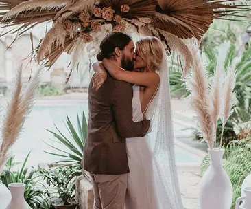 A bride and groom share a kiss under a bohemian floral arch at their outdoor wedding ceremony.